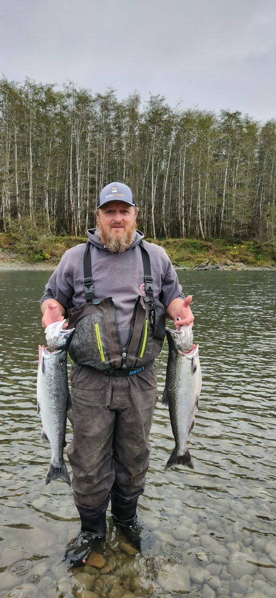 An angler in waders standing in the river holding a salmon in each hand, bare alder trees behind