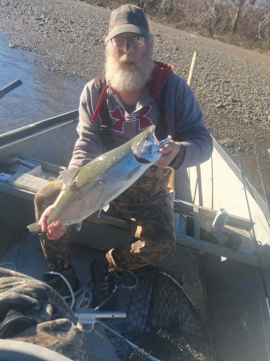 An angler kneeling in the drift boat holding a bright salmon, gravel bar behind