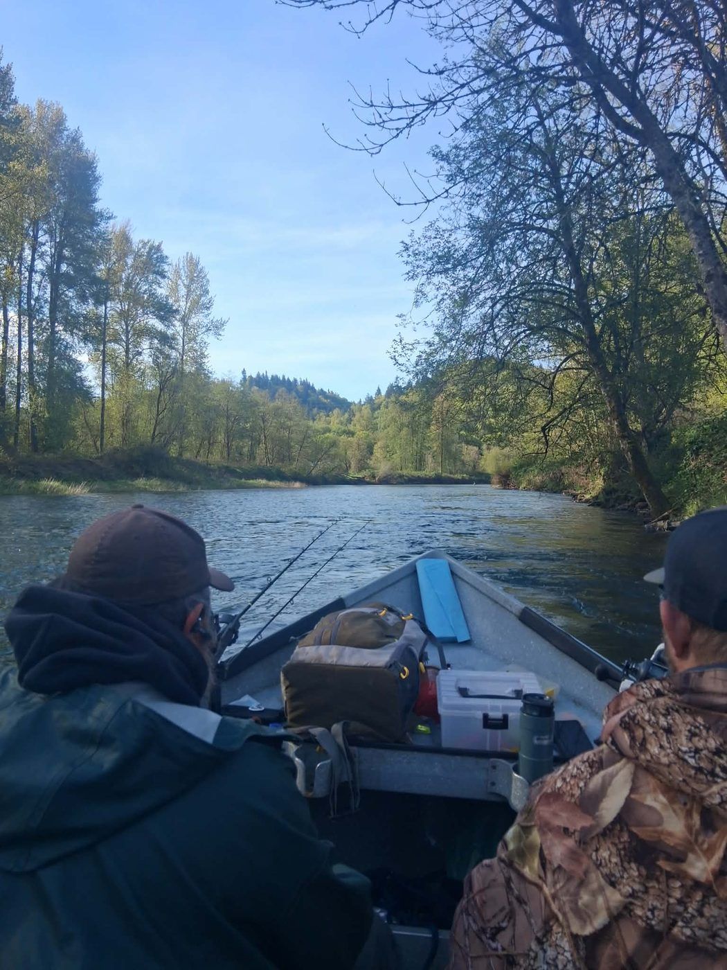 Two anglers seated in a drift boat working their rods as the boat drifts a forested stretch of river